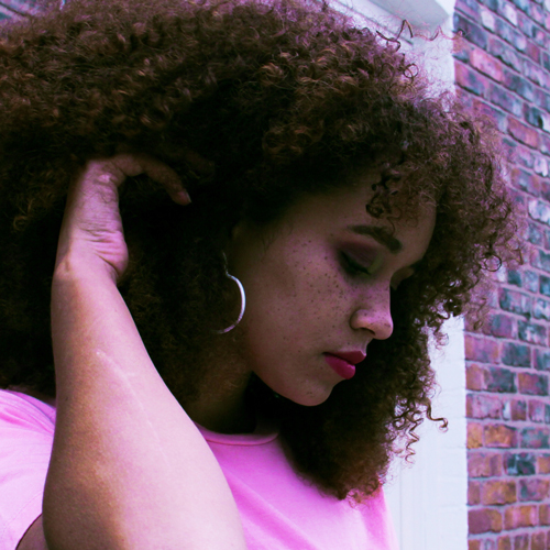 Girl with curly, brown hair and a pink T-shirt is putting her hair behind her ear and looking down at the floor.