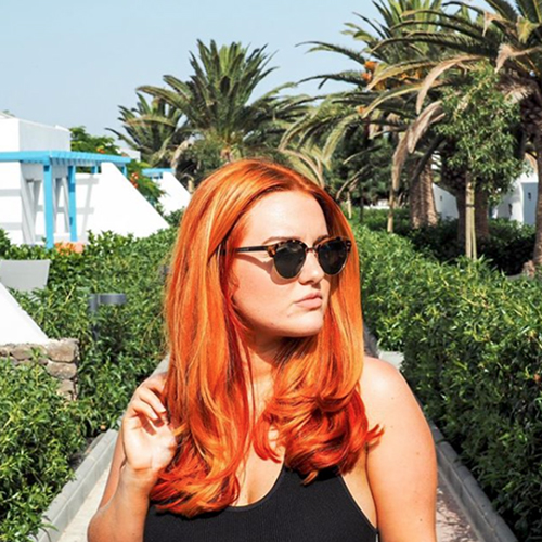 Girl with wavy, red hair is standing in a park and wearing a black string top and sunglasses while looking away from the camera.
