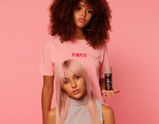 Girl with straight, pink hair is sitting in front of a girl with curly, brown hair who is standing up and holding the Restora Protein Conditioner in the palm of her left hand.