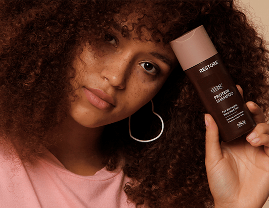 Girl with curly, brown hair holds the Restora Protein Shampoo against her forehead.