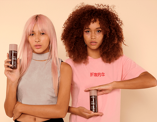 Girl with straight, pink hair holds the Restora Protein Shampoo in her left hand and is standing beside a girl with curly, brown hair who holds the bottle between her hands.