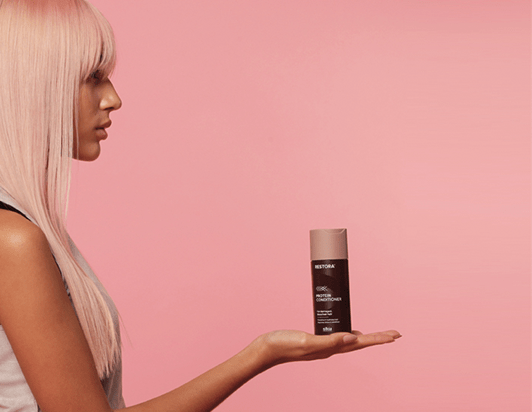 Girl with straight, pink hair is turned to the side and holds the Restora Conditioner in the palm of her right hand.